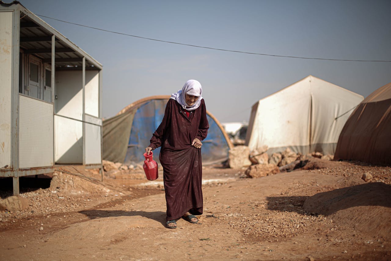 services-01 An elderly woman walking in a Syrian refugee camp, carrying a red container.